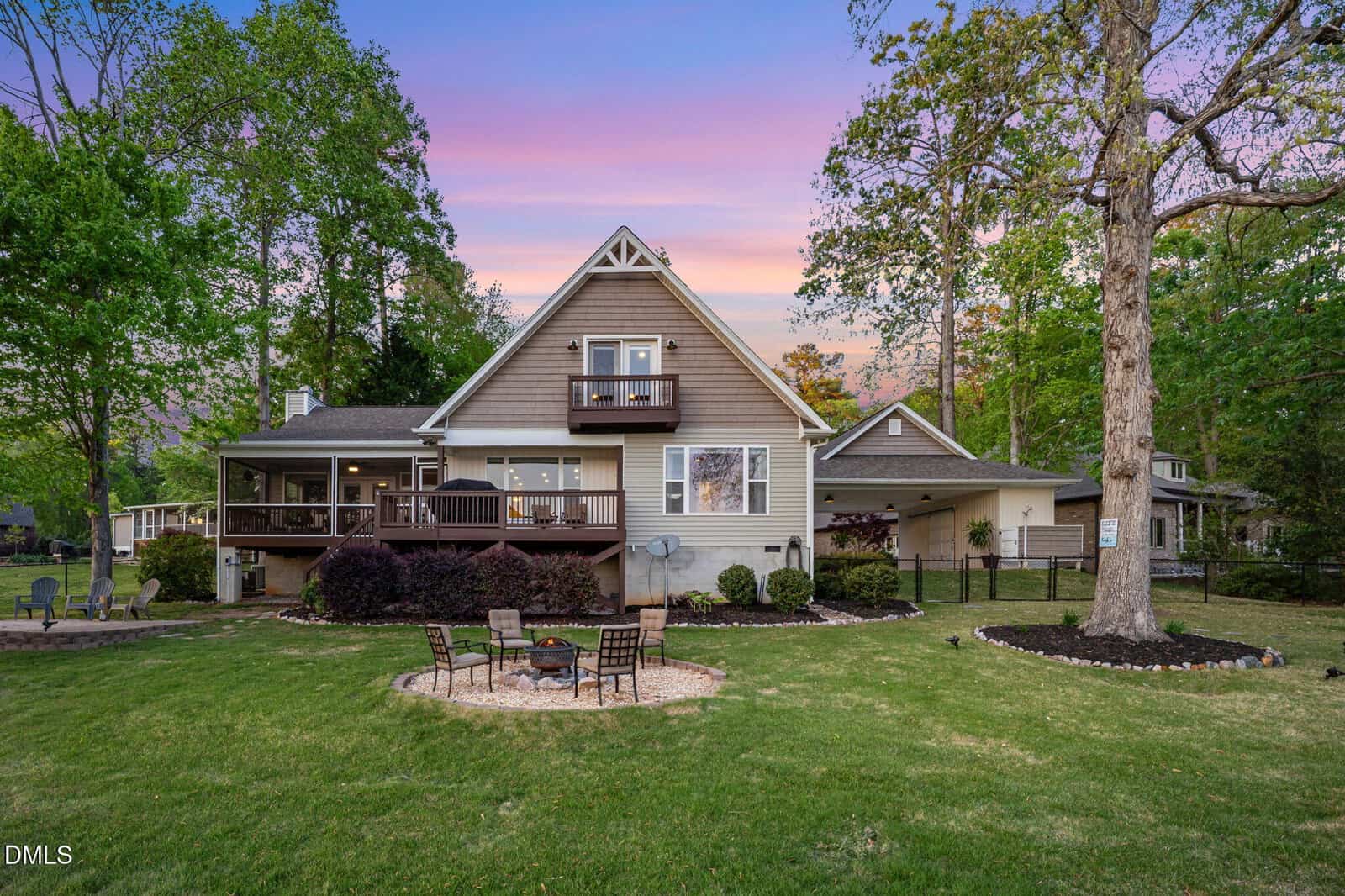 A two-story house with a small upper balcony, large covered deck, and landscaped backyard featuring a fire pit with chairs. The scene is set at sunset with trees and neighboring houses visible.