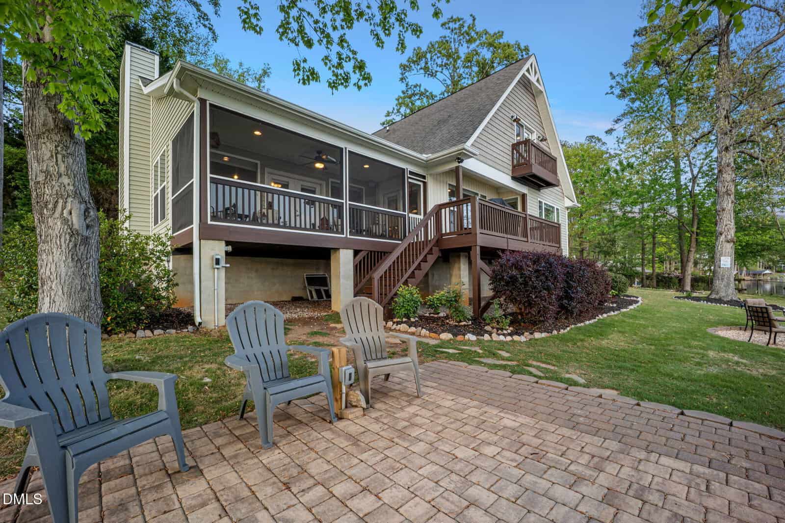 A modern lake house with a screened porch, balcony, and wooden stairs, surrounded by trees. Four plastic chairs sit on a paved patio facing the house. A grassy lawn leads to the water in the background.
