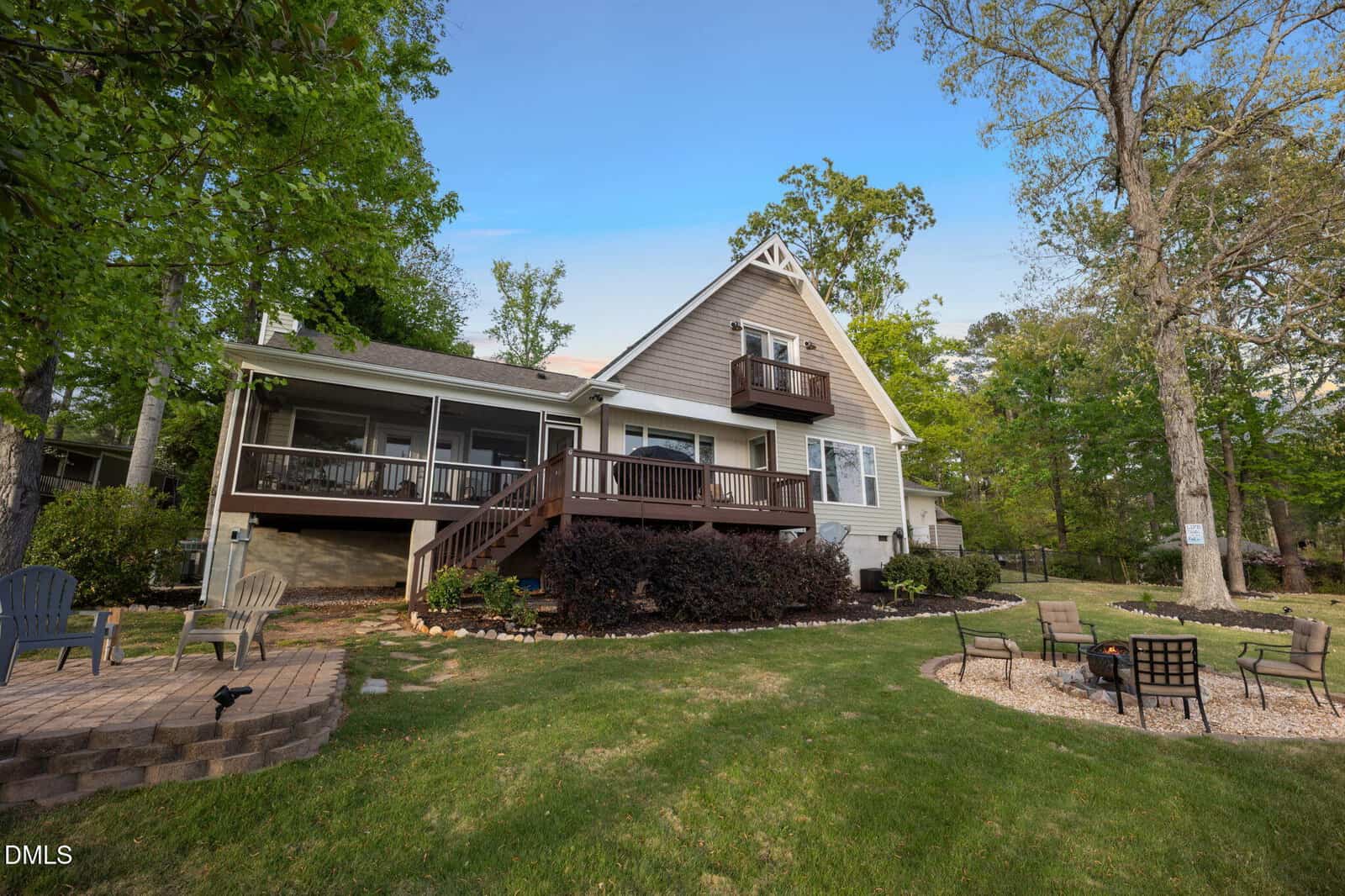A two-story house with a wood deck, screened porch, and small balcony, surrounded by trees, green lawn, and outdoor seating areas under a blue sky.