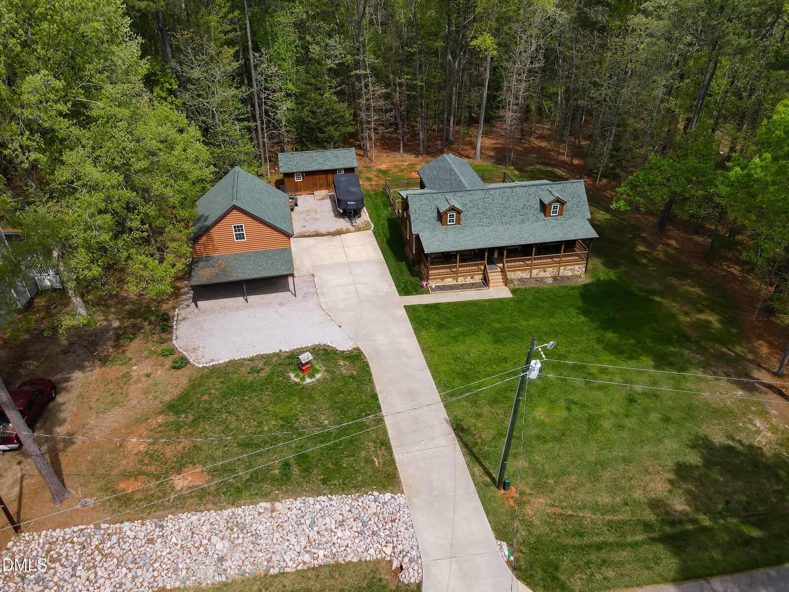 Aerial view of a wooded property with a log-style house, detached garage, and long driveway. The house has a front porch and green roof, surrounded by grass and trees. A few vehicles and a gravel area are visible.
