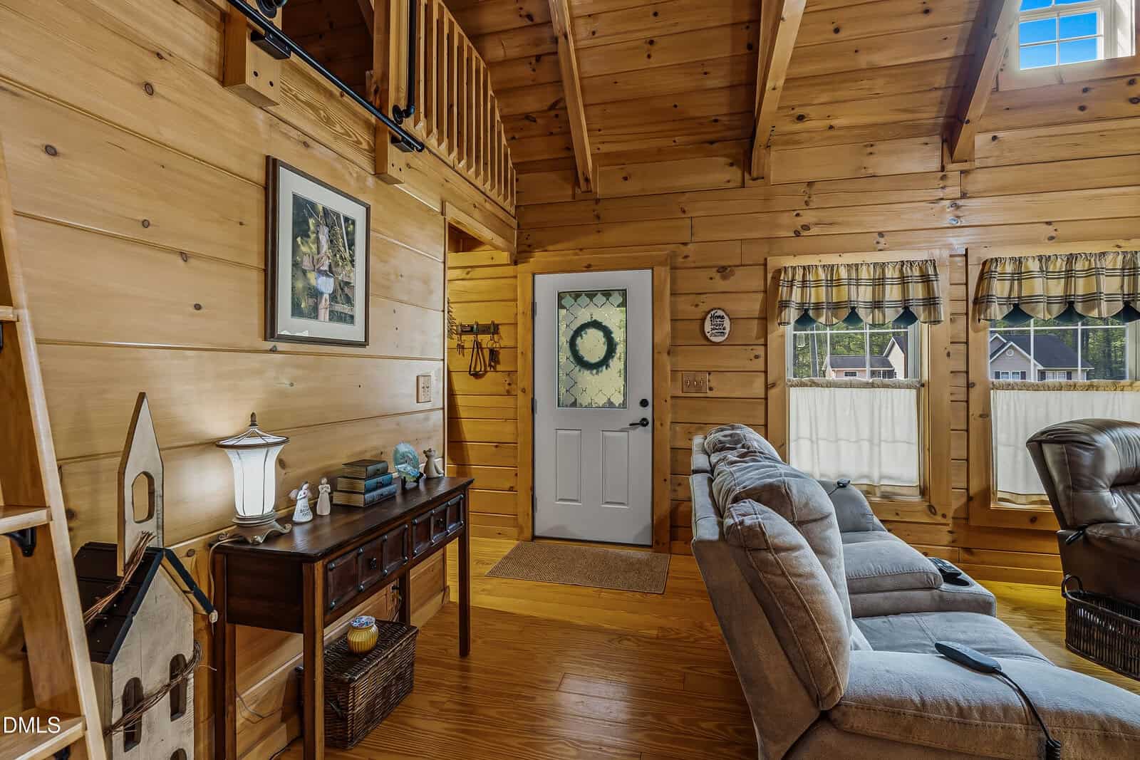 A cozy living room with wood-paneled walls and ceiling, gray recliner chairs, a wooden console table with a lamp and decor, and a door with a wreath. Two windows with plaid curtains let in natural light.