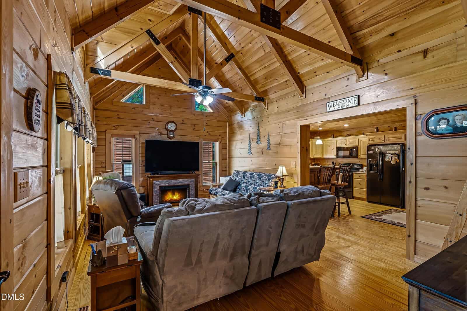 A cozy log cabin living room with wood-paneled walls, vaulted ceiling, gray sofas, a fireplace, and a TV. The open kitchen and dining area are visible in the background. Ceiling fan and rustic decor add warmth.
