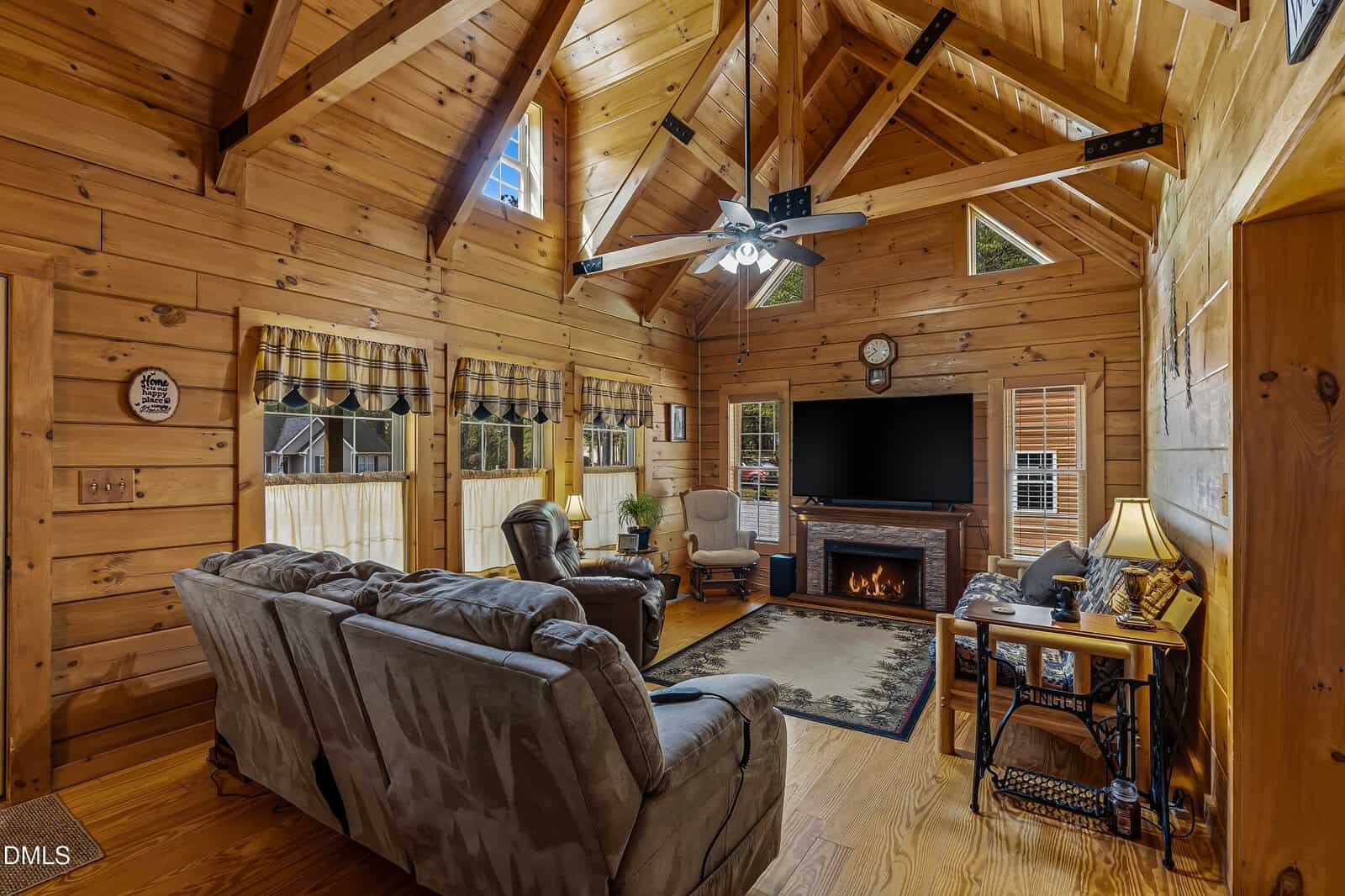 A cozy log cabin living room with wood walls, vaulted ceiling, large windows, a sectional sofa, armchairs, a TV above a fireplace, and a ceiling fan with lights. Natural light brightens the warm, rustic space.