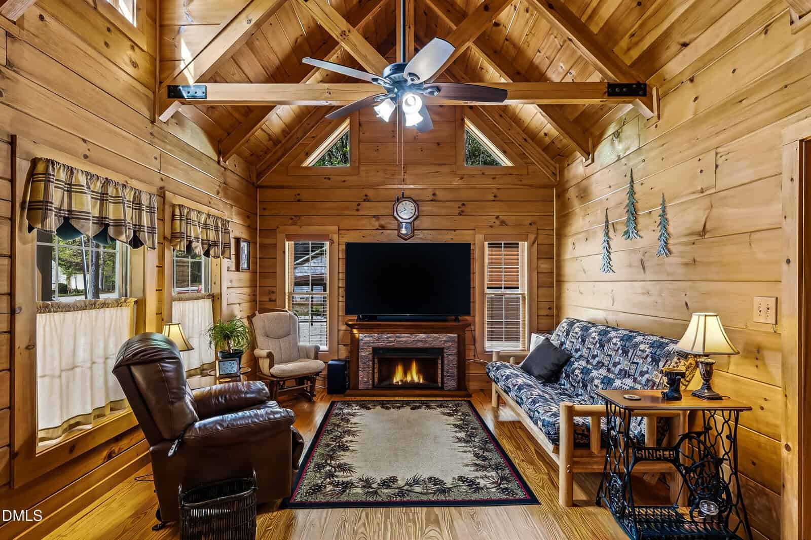 Cozy wooden cabin living room with TV, fireplace, patterned sofa, recliner, armchair, lamps, and windows with plaid valances. Ceiling fan above and decorative greenery on the wall.