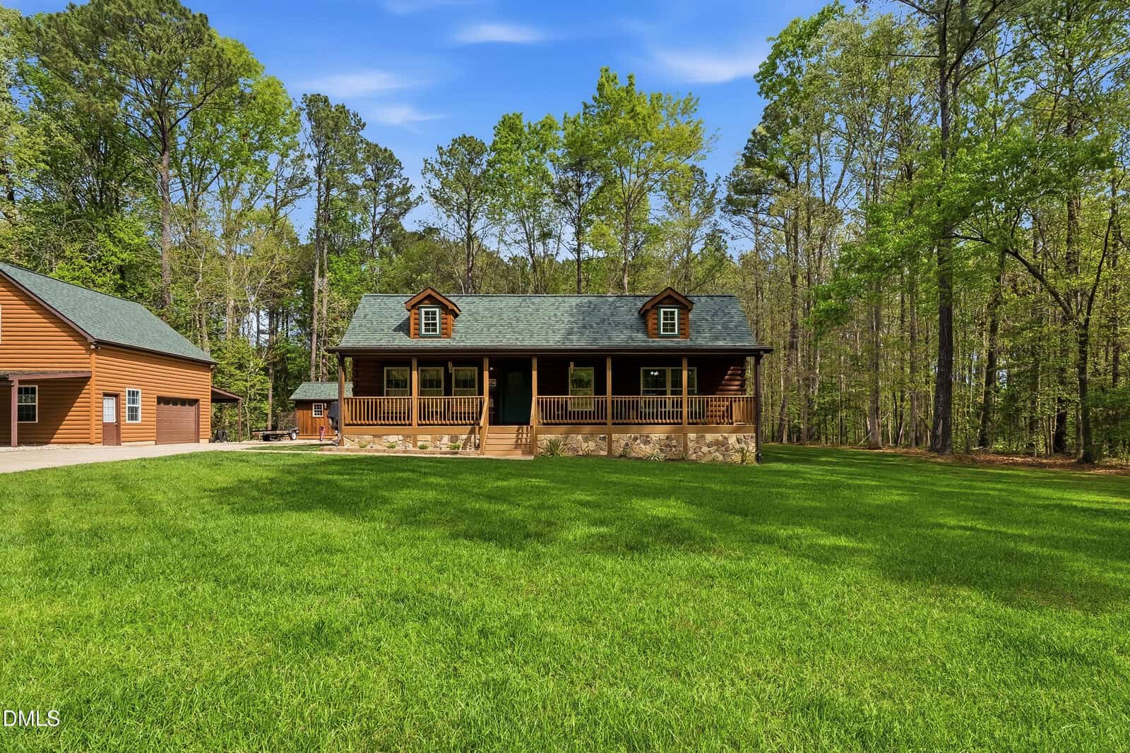 A wooden cabin-style house with a front porch sits on a large, green lawn, surrounded by tall trees. An attached garage is visible on the left. The sky is clear and blue.