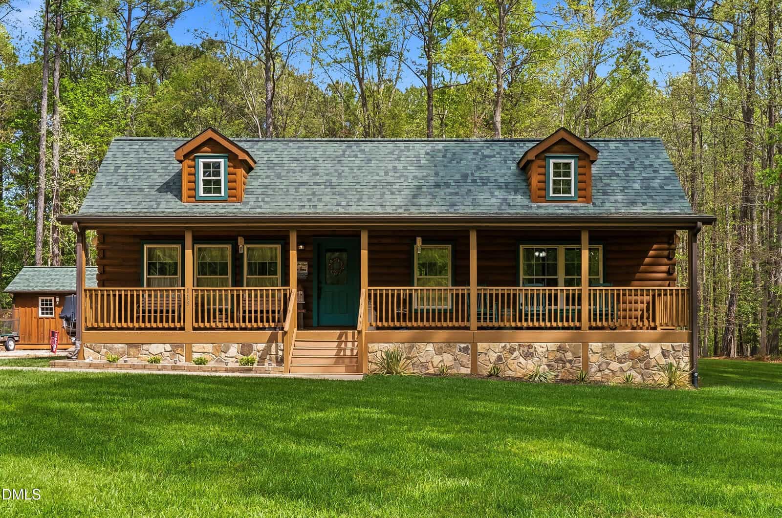 A cozy log cabin at 115 Running Bear Drive, Louisburg, with a green roof and two dormer windows, features a covered front porch with wooden railings, set on a lush green lawn and surrounded by tall trees.