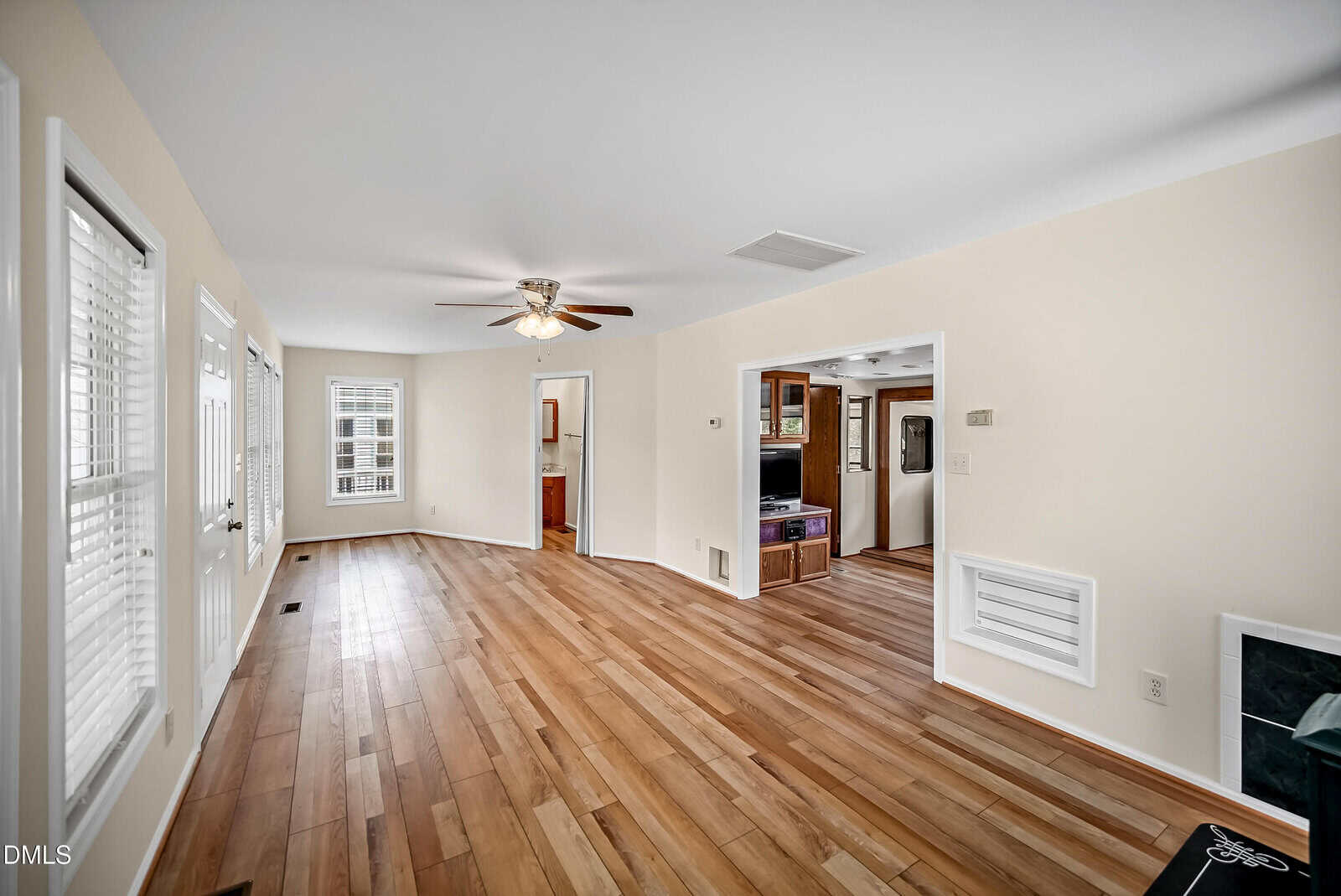Bright living room with light wood floors, beige walls, ceiling fan, multiple windows, and open entryways leading to other rooms. A small vent and thermostat are visible on the right wall.