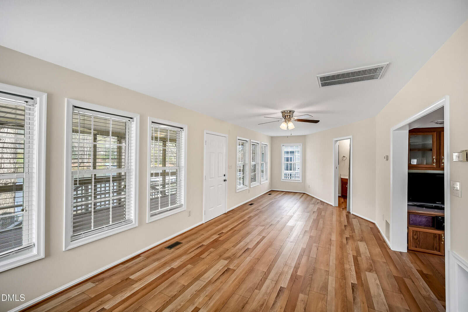 Spacious, bright room with light wood flooring, beige walls, multiple windows, ceiling fan, and white trim. Doorways lead to other rooms; porch visible through windows.