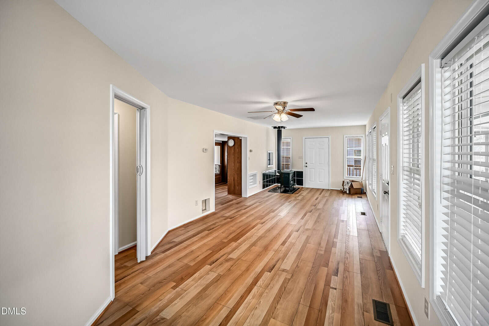 A spacious room with light wood floors, beige walls, white trim, many windows, a ceiling fan, a black wood stove, and multiple doorways leading to other areas.