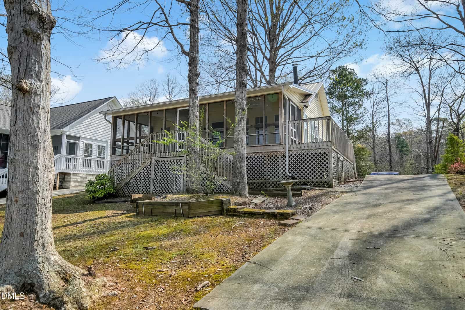 A wooden house with a screened-in porch sits on a raised foundation next to a concrete driveway, surrounded by bare trees and a patchy lawn under a partly cloudy sky.