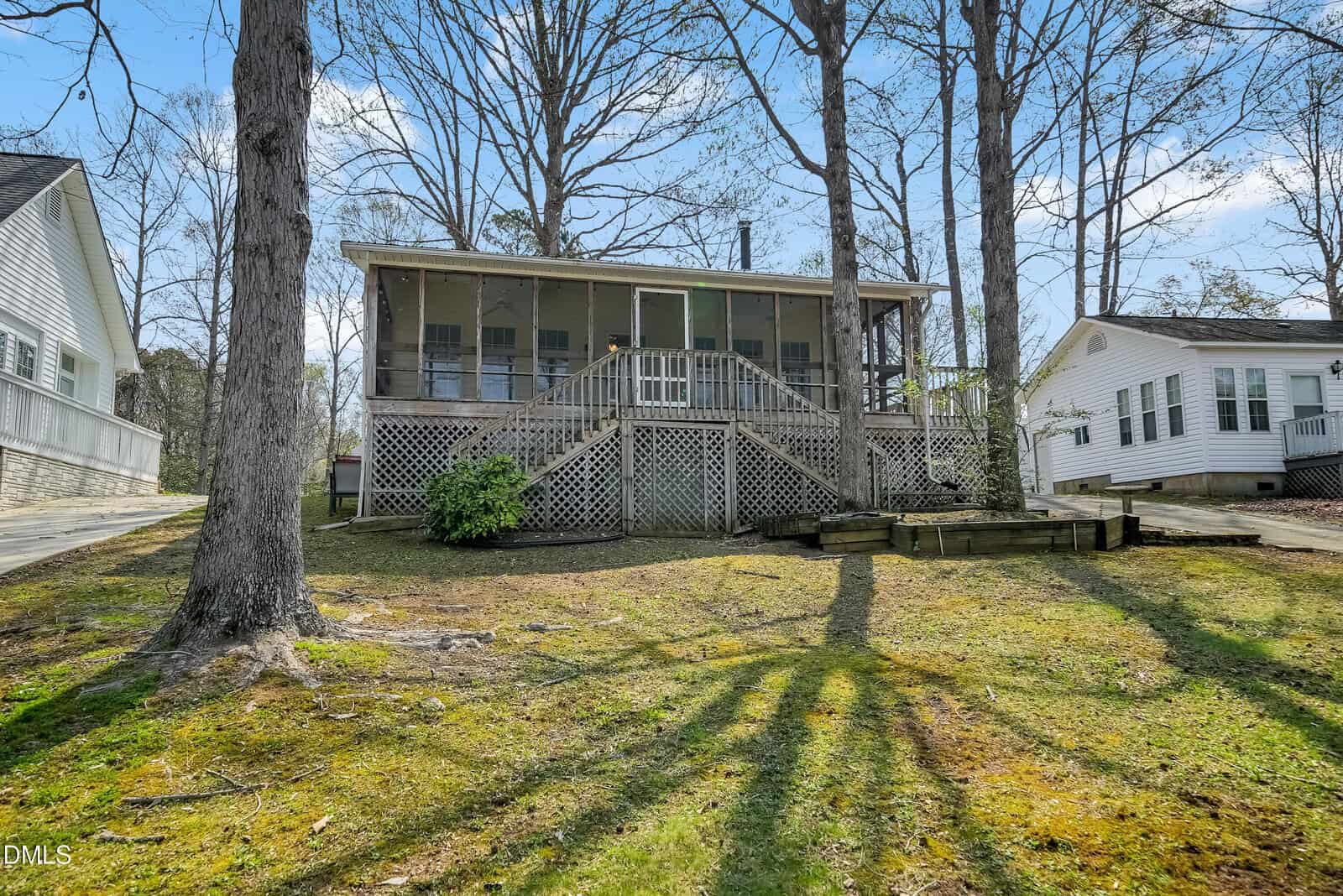 A wooden house with a large screened porch sits on a sloped, grassy yard. Wooden steps lead up to the porch. Neighboring houses are visible on both sides, and tall trees surround the homes under a partly cloudy sky.
