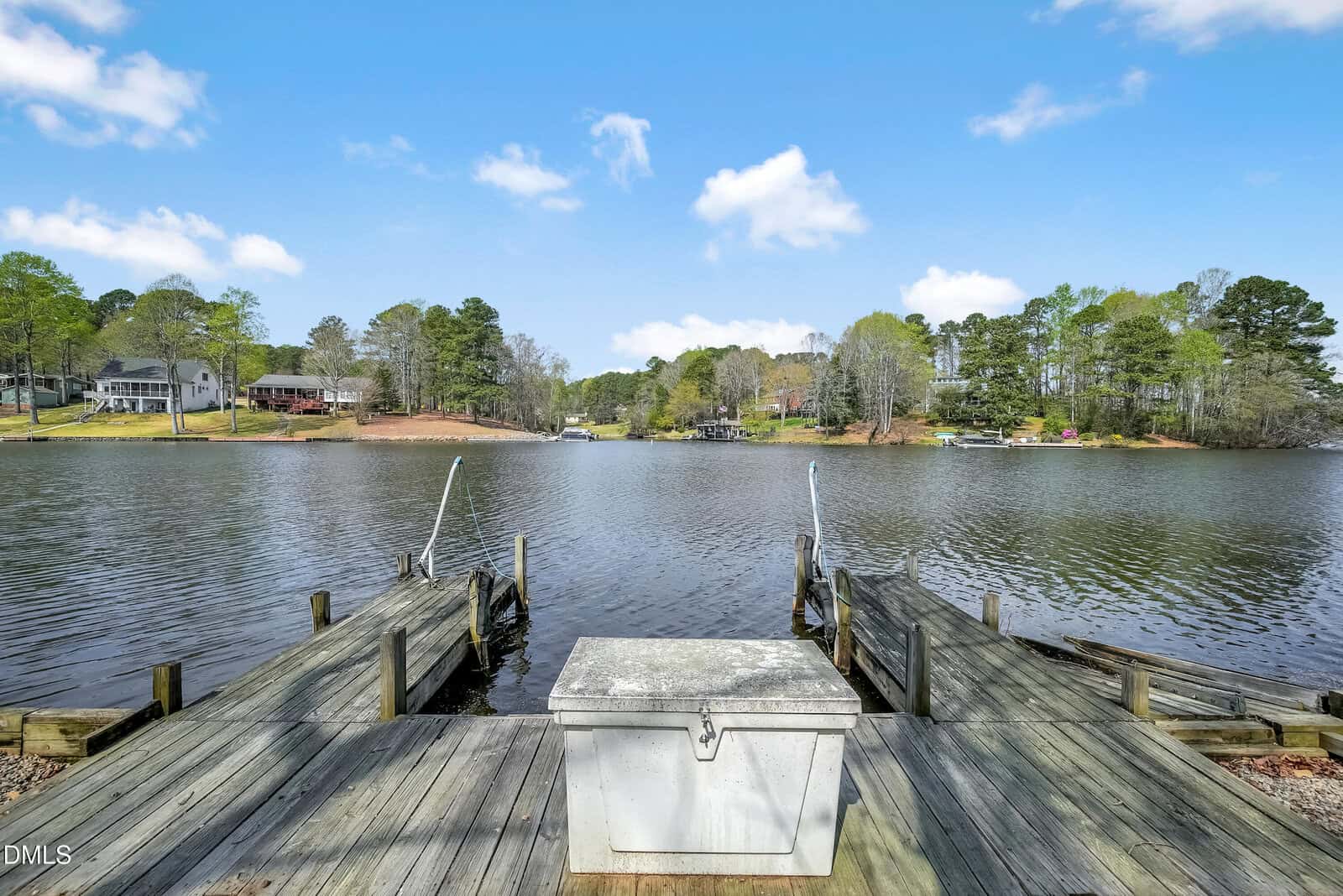 A wooden dock with a metal storage box at the end extends into a calm lake, surrounded by trees and houses under a blue sky with scattered clouds.