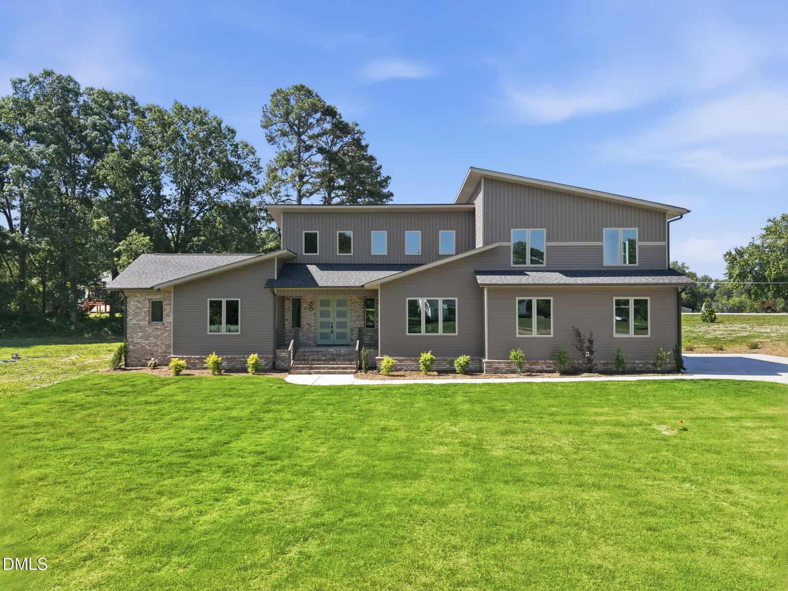 Modern two-story house with gray siding, large windows, and a double front door. The home sits on a well-manicured lawn with small shrubs and trees, under a clear blue sky.
