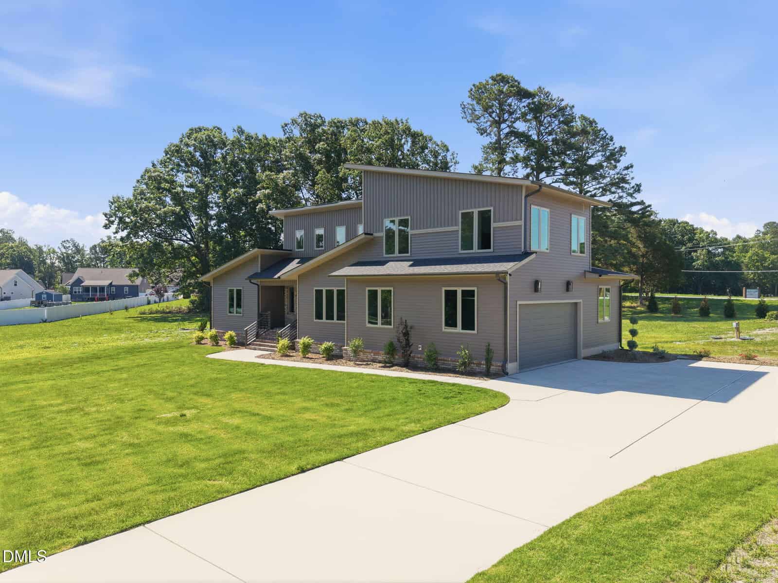 Modern two-story house at 50 Wiggins Road, Louisburg, with light gray siding, large windows, and an attached garage, set on a spacious green lawn with a long driveway and surrounded by trees under a clear blue sky.