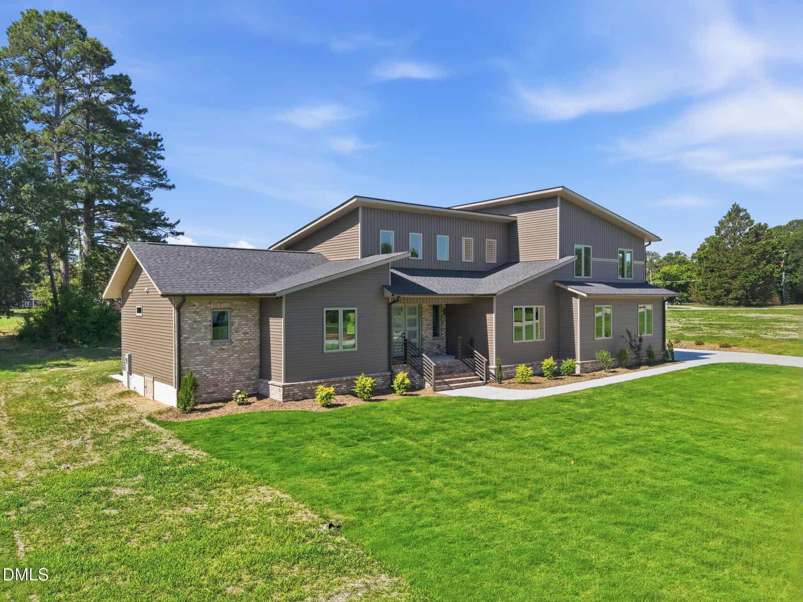 Modern two-story house with brown siding and stone accents, large windows, and a dark shingle roof, set on a spacious green lawn with a few trees in the background under a blue sky.