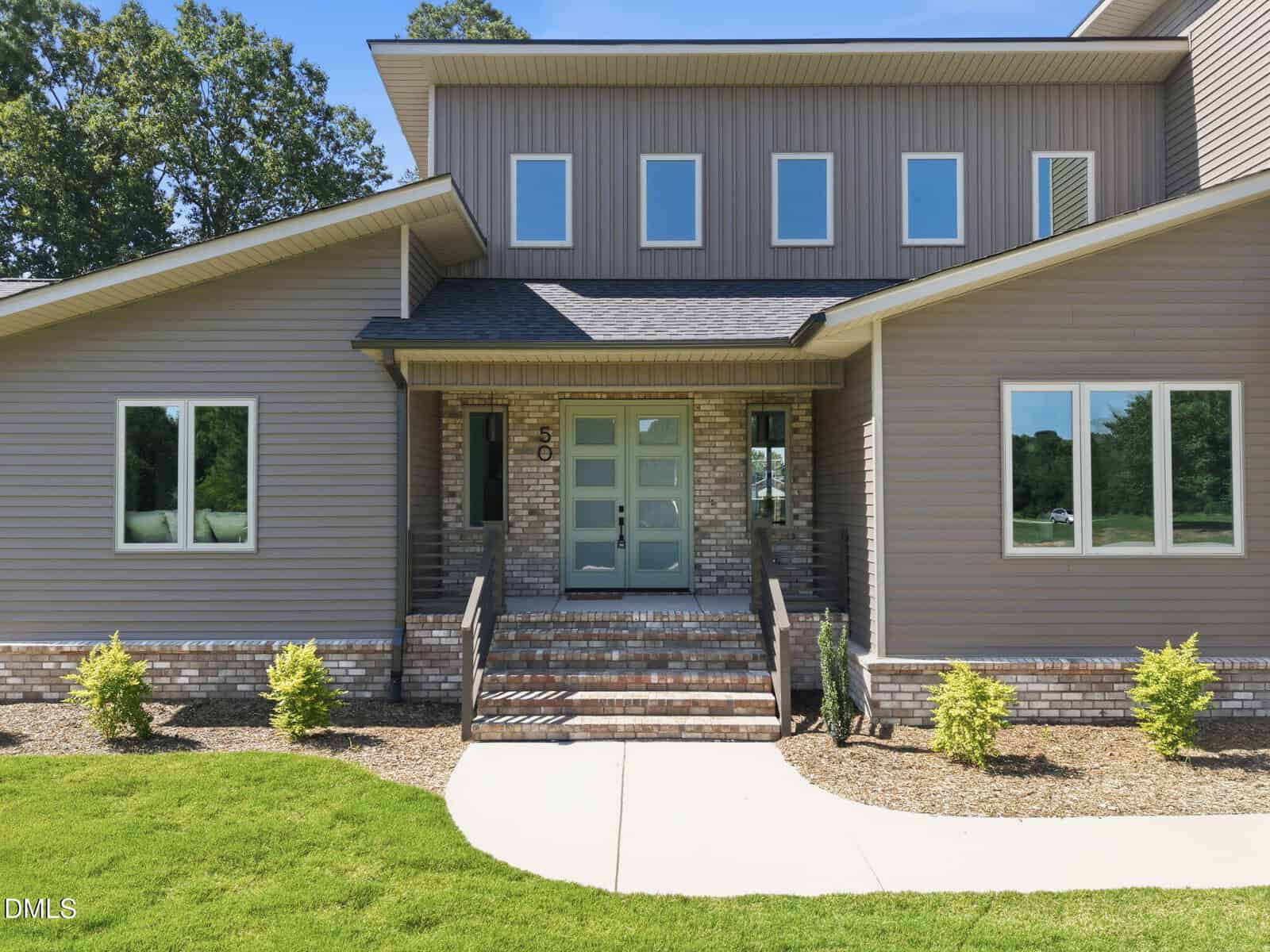 Modern two-story house with gray siding, brick accents, and multiple rectangular windows. The entry features double glass doors, steps, and a landscaped front yard with small shrubs and green grass.