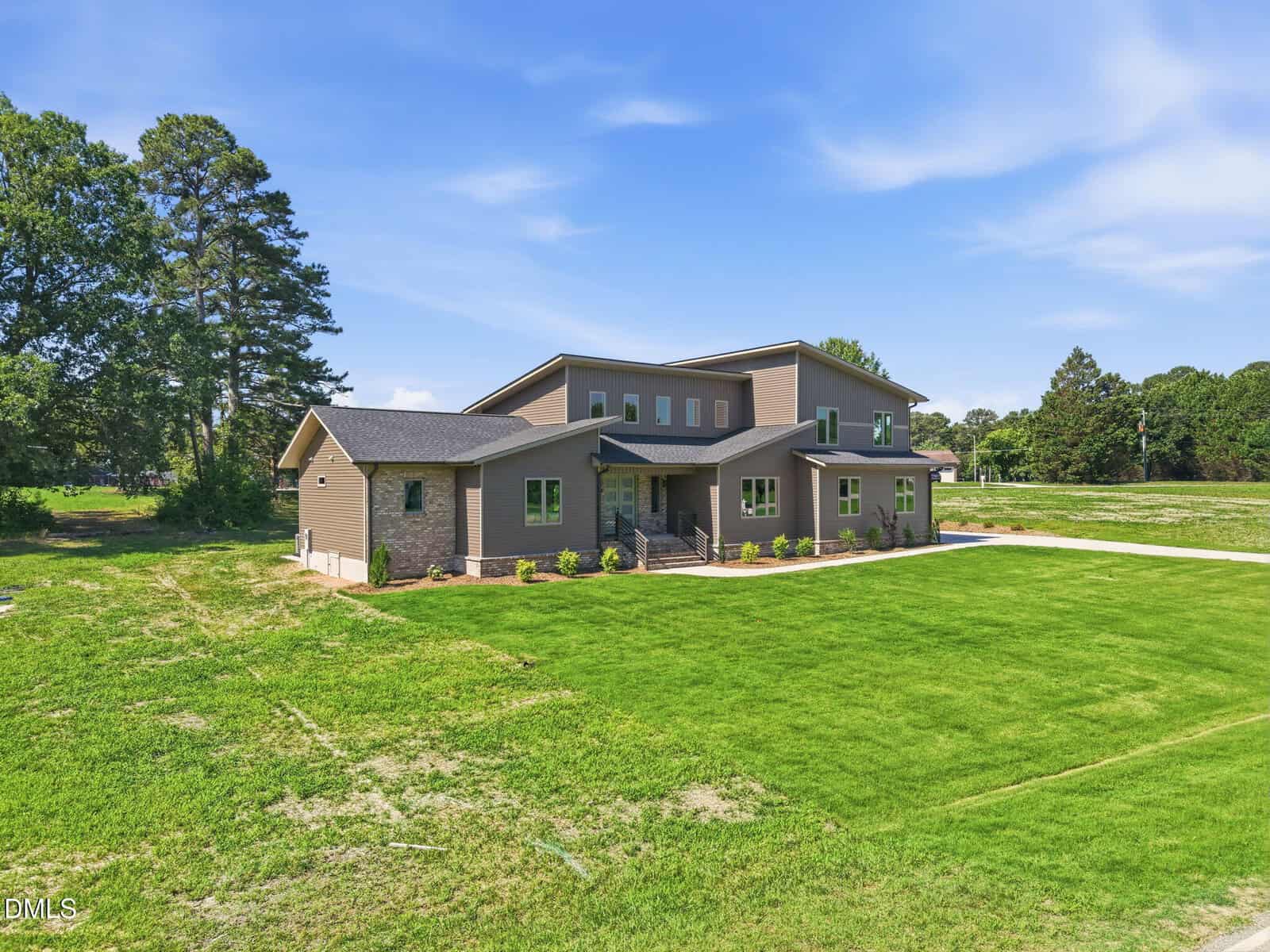 Modern two-story house with brown siding, large windows, and a brick accent foundation, surrounded by a spacious, well-maintained lawn and bordered by mature trees under a clear blue sky.