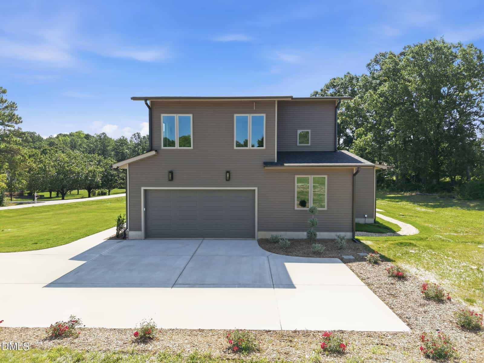 A modern two-story house with gray siding, double garage doors, and large windows, surrounded by a wide driveway, small shrubs, and green lawns with trees in the background under a blue sky.