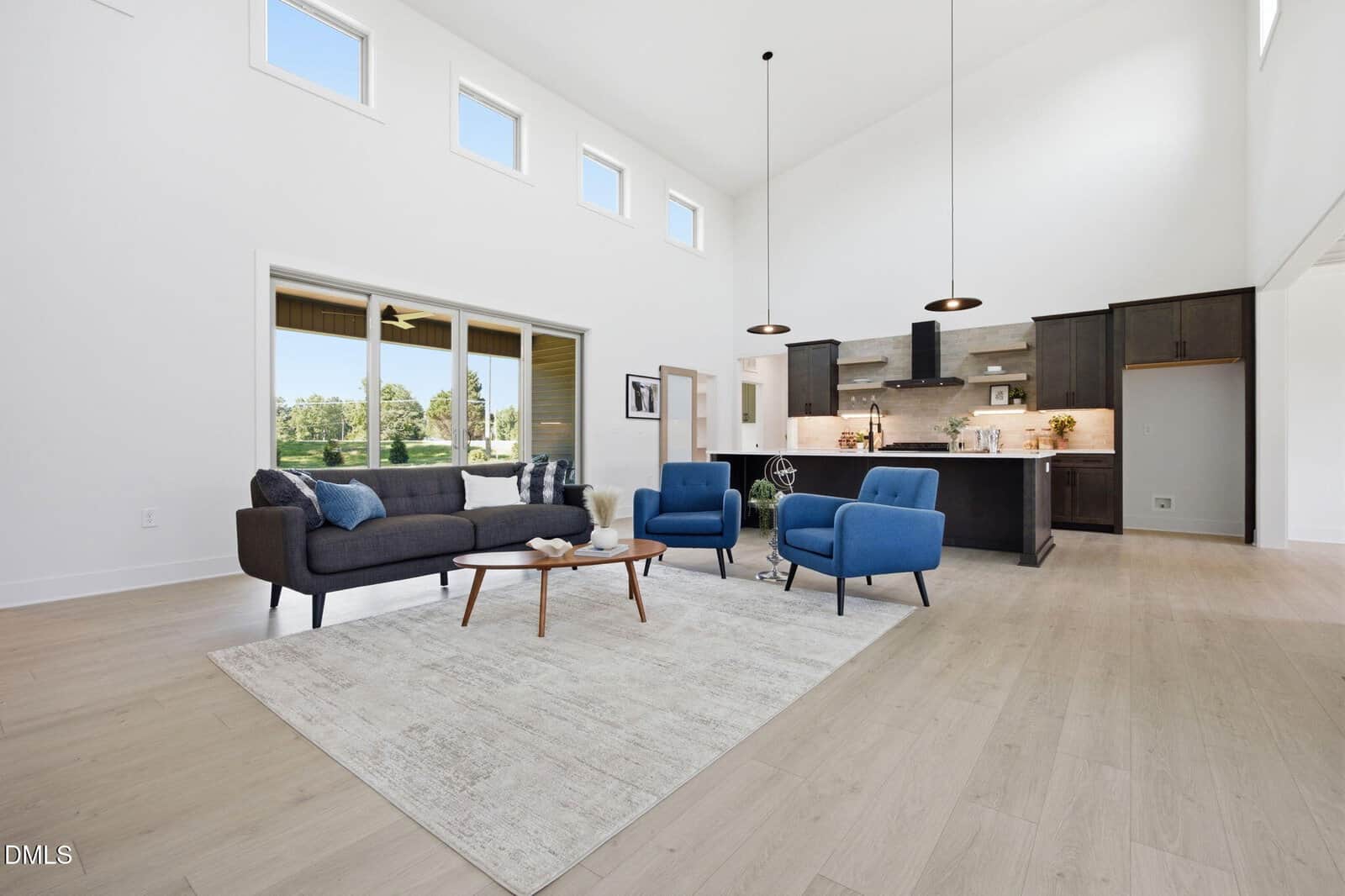 Modern living room with light wood floors, white walls, blue armchairs, a dark sofa, a beige rug, and a coffee table. Open kitchen in the background with pendant lights and large windows letting in natural light.