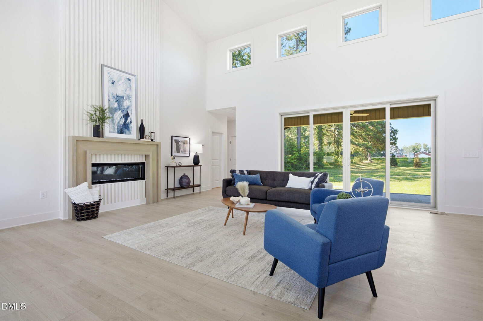 Bright living room with tall white walls, large windows, a modern fireplace, gray sofa, blue armchair, light wood floors, a rug, and sliding glass doors opening to a green yard.