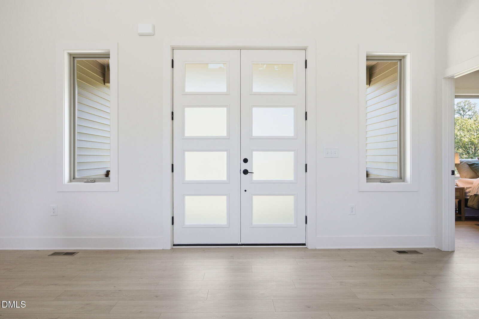 A modern white front door with six frosted glass panels is centered between two rectangular windows. The room has light wood flooring and white walls, creating a bright, minimalist look.
