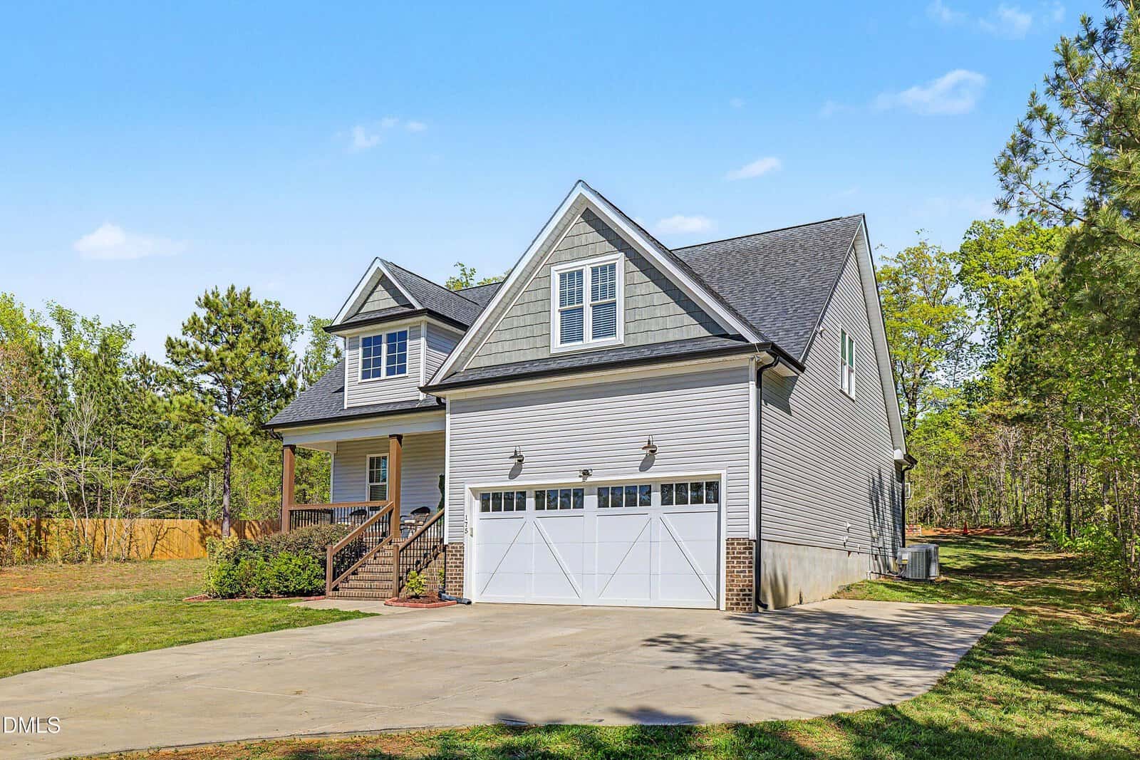 A two-story gray house with white trim and a large attached garage sits on a spacious lot with a concrete driveway, surrounded by trees and grass under a clear blue sky.