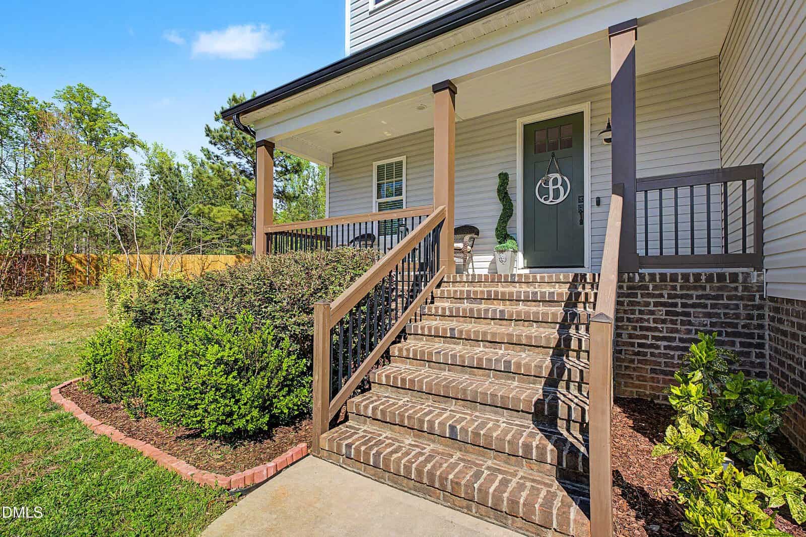 A brick staircase with black railings leads to a covered porch with two chairs, potted plants, and a green door displaying a circular monogram. Shrubs and mulch border the walkway and house.
