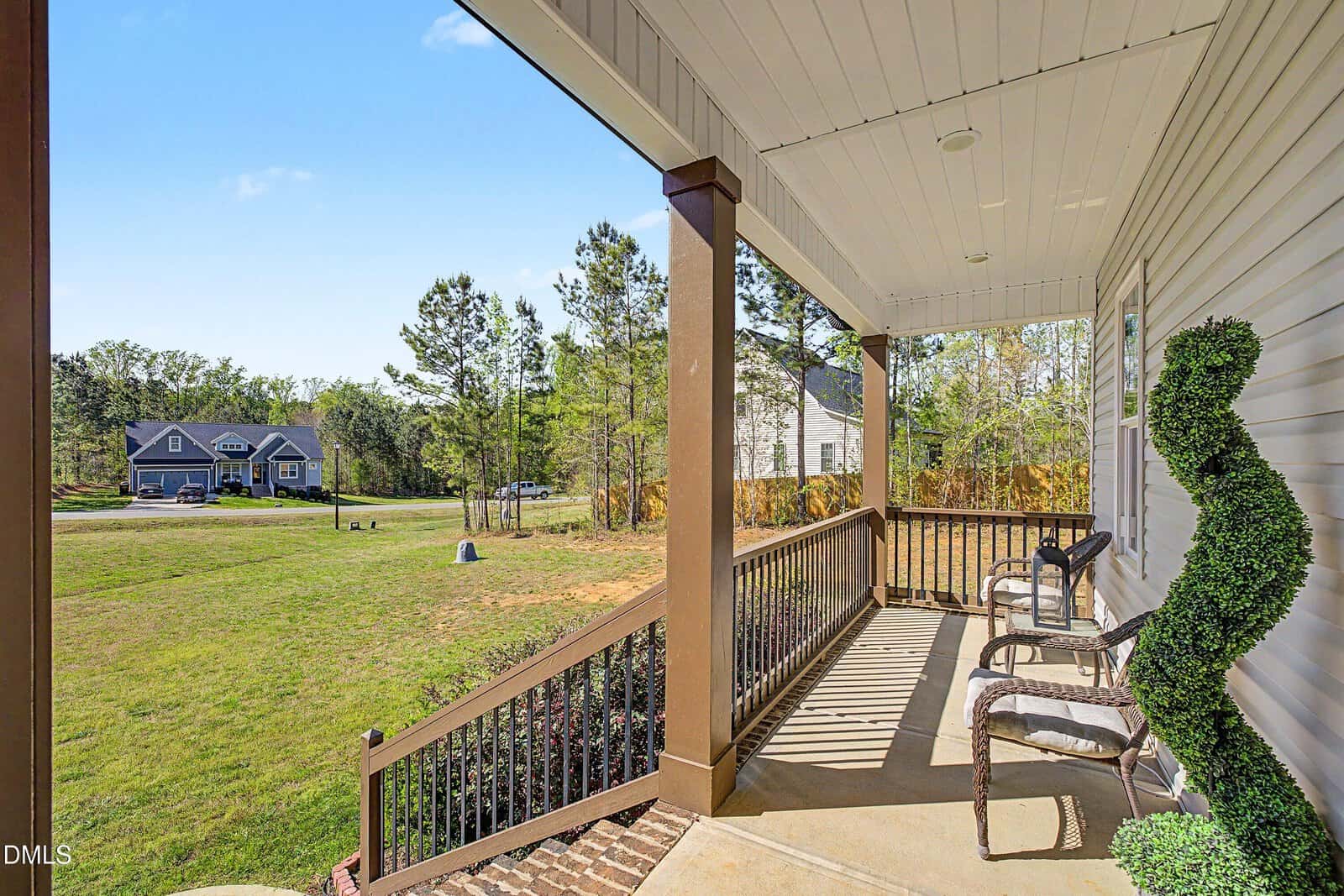 A covered porch with two chairs and a topiary overlooks a grassy yard. Trees border the property, and houses are visible in the distance under a blue sky. Shadows from the railing fall across the porch floor.
