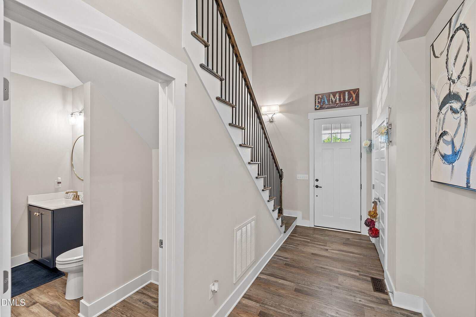Bright entryway with wood flooring, white walls, staircase with dark railing, front door with window, “FAMILY” sign above, a bathroom on the left, modern decor, and a colorful abstract painting on the right wall.