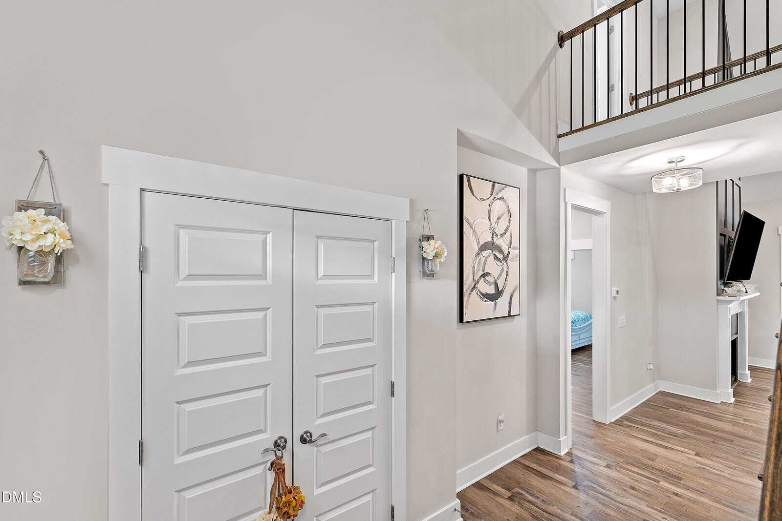 Bright hallway with double white doors, hardwood floors, wall art, and a view into another room. A railing and loft are visible above, and small flower decorations hang on the wall near the door.