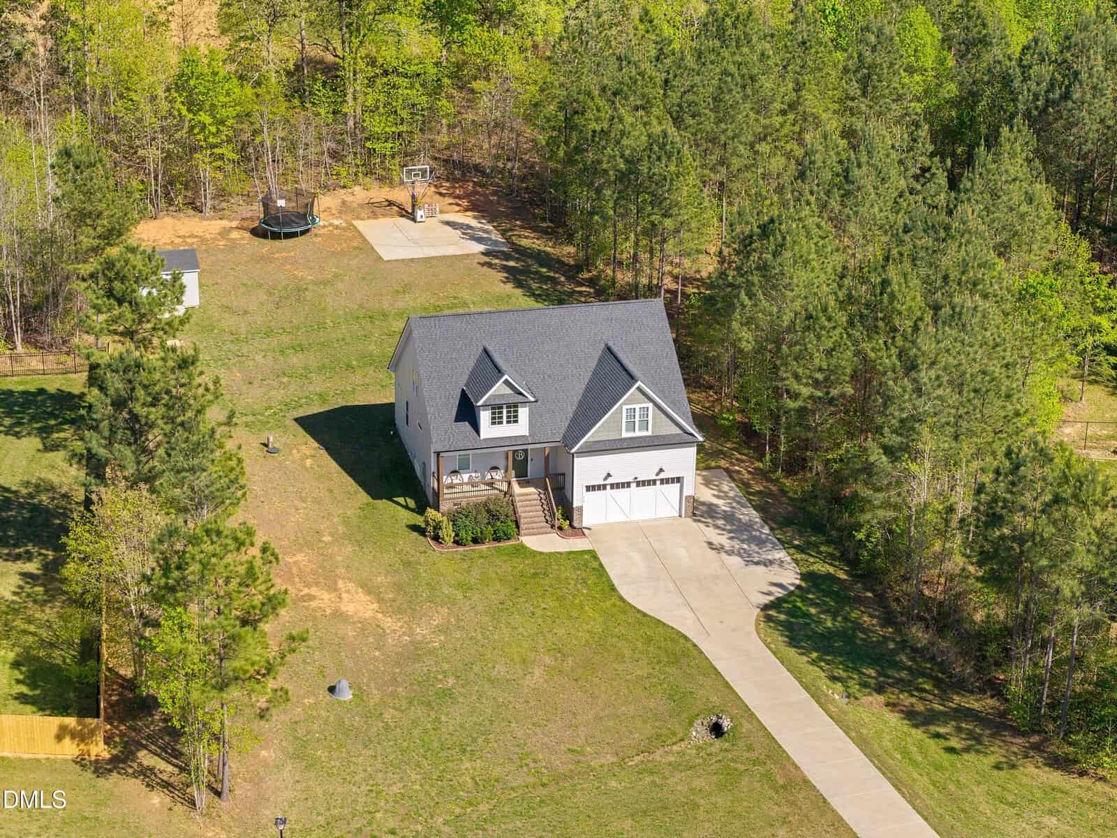 A two-story house with a gray roof sits on a large green lot, surrounded by trees. A long driveway leads to a three-car garage. A trampoline and basketball hoop are visible in the backyard.