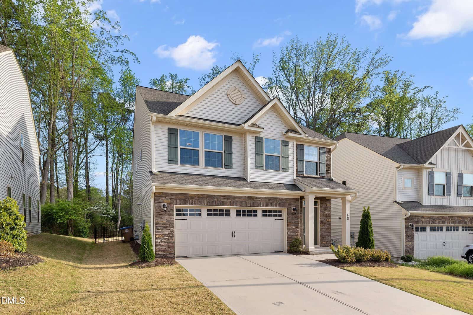 Two-story beige house with a double garage, brick accents, and a neatly landscaped yard. Surrounded by trees and neighboring homes, with a clear blue sky above.