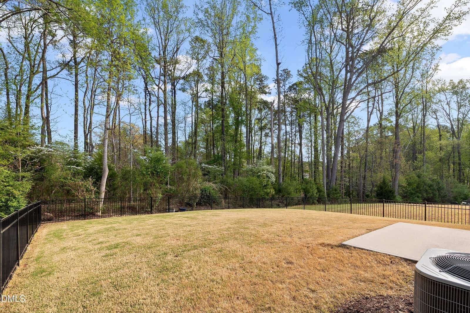 Fenced backyard with a grassy lawn, some brown patches, a small concrete patio, and tall trees in the background under a blue sky with scattered clouds.