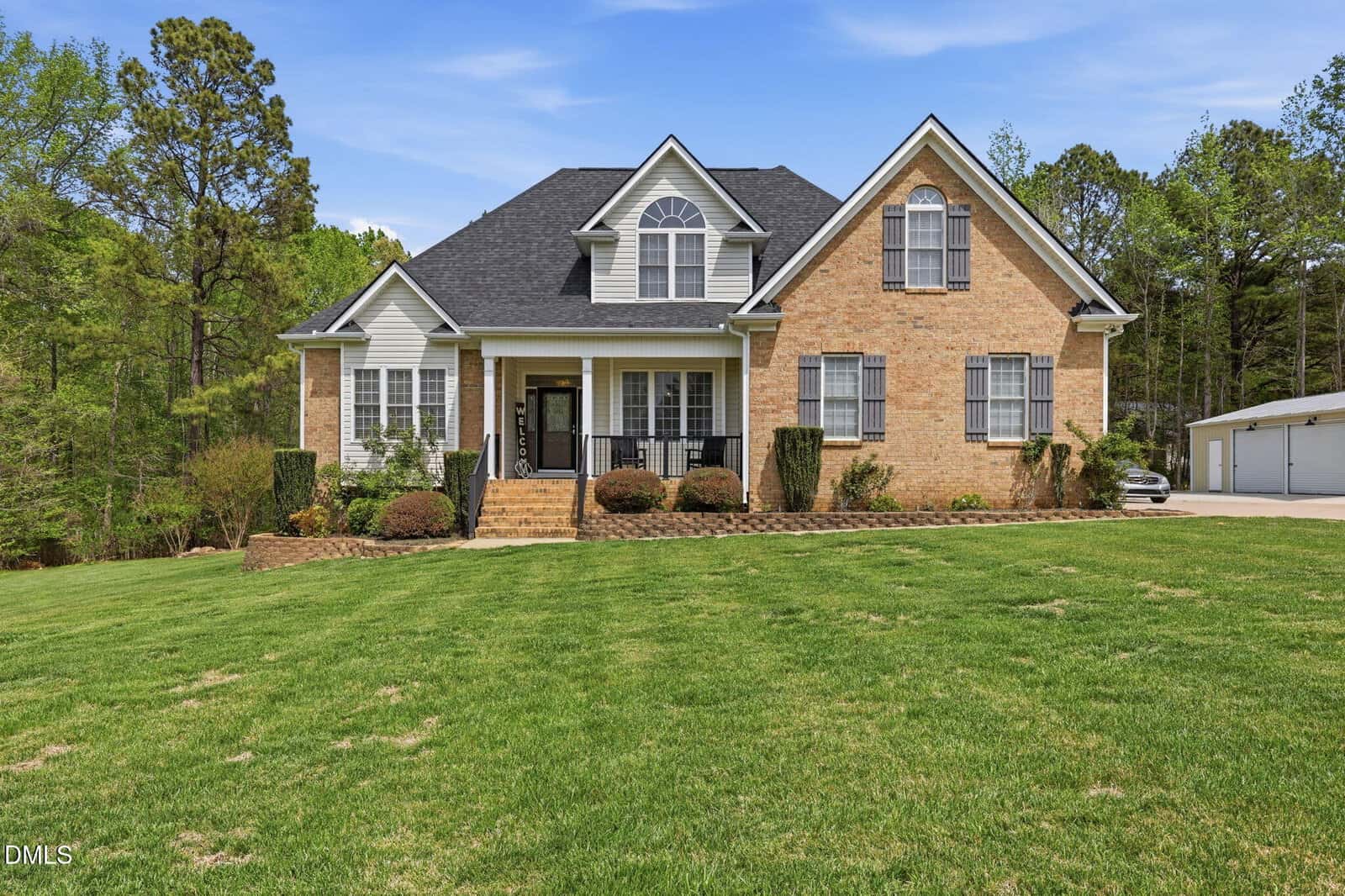 Two-story brick house with dark roof, black shutters, and front porch, set on a large grassy lawn with shrubs. Trees and a driveway with a garage are visible in the background under a blue sky.