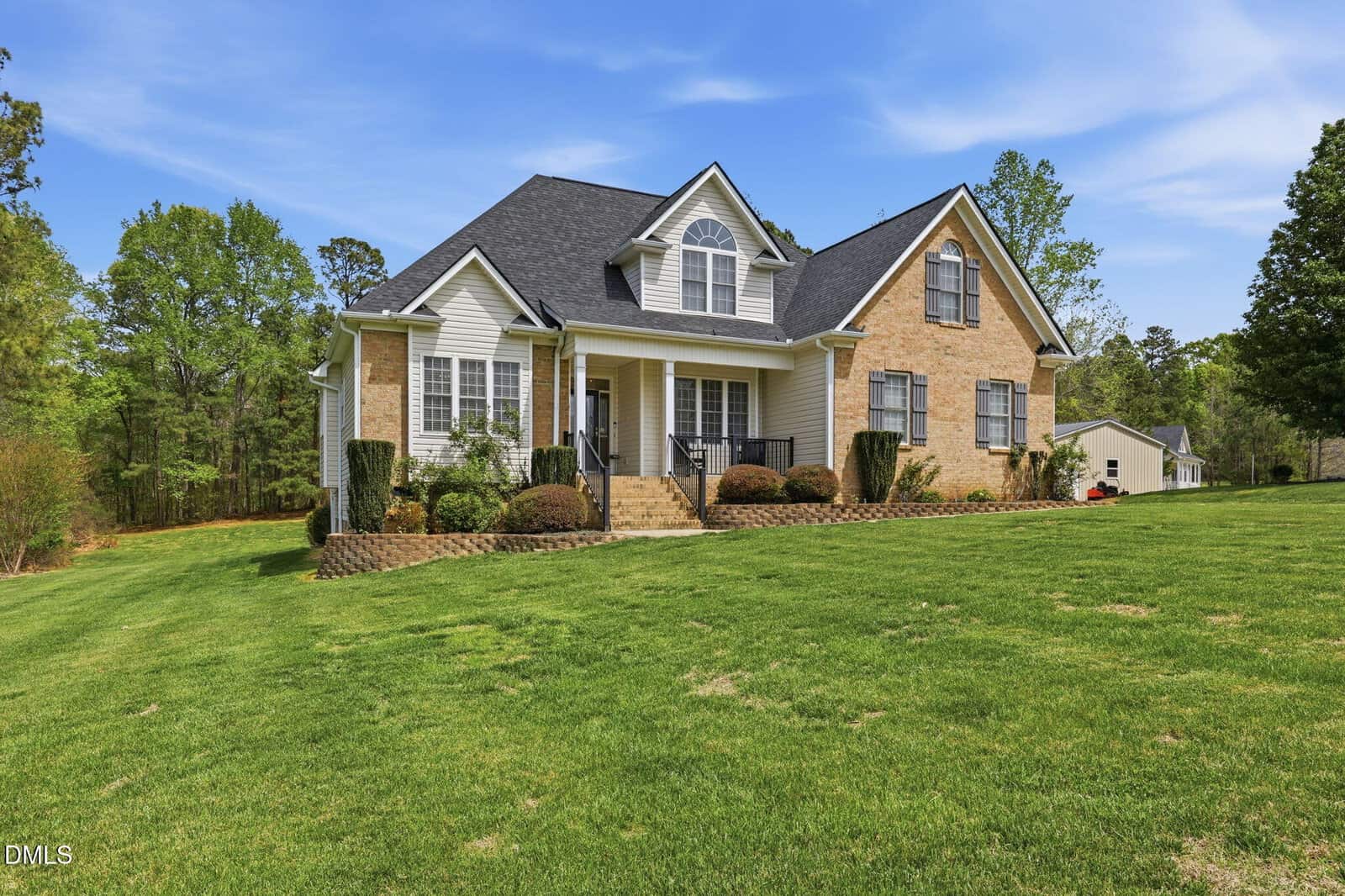 Two-story brick and siding house with gabled roof, front porch, steps, manicured bushes, and large green lawn, surrounded by trees under a blue sky.