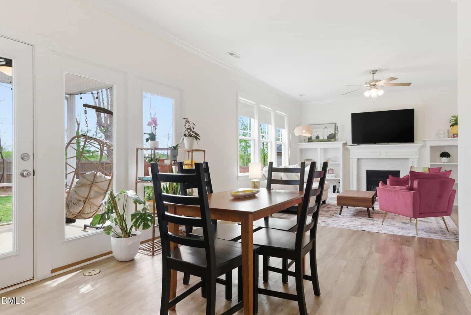 Bright dining and living area with wood floors, black dining chairs, a fireplace, a TV, pink armchairs, plants, and large windows letting in natural light.