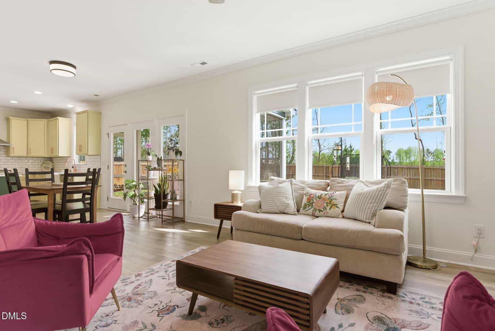 Bright living room with a beige sofa, pink armchairs, wooden coffee table, floor lamp, and floral rug. Large windows let in natural light. Dining area and kitchen are visible in the background.