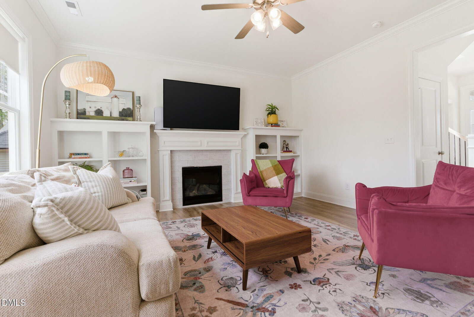 Bright living room with a beige sofa, two pink armchairs, a wooden coffee table, floral rug, and a fireplace with a large TV above it; shelves hold decor and plants, and a floor lamp stands by the window.