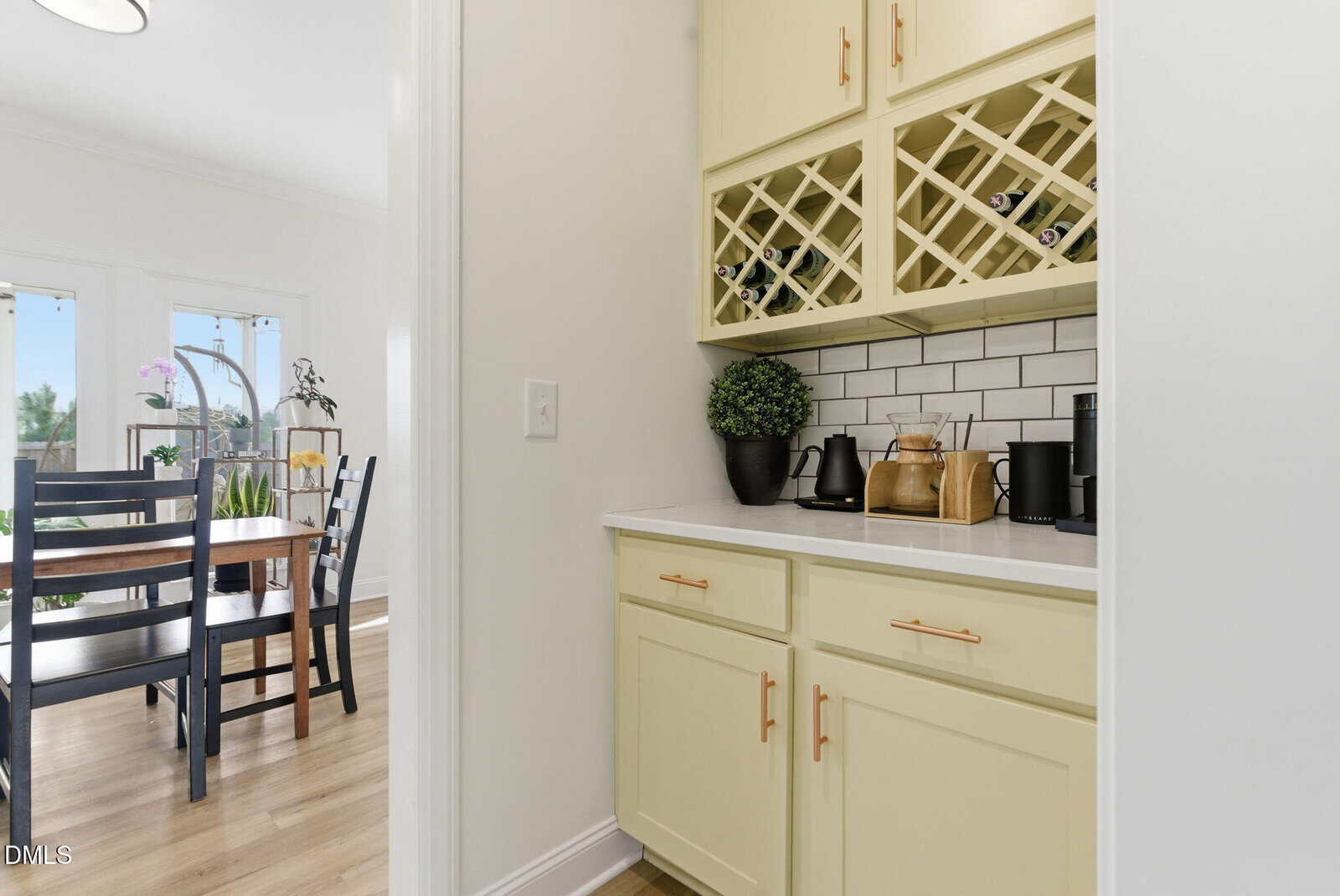 A cream-colored wet bar features a wine rack filled with bottles, a coffee maker, and a small plant, next to a dining area with a wooden table and chairs.