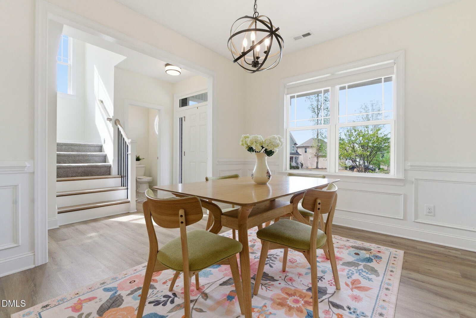 A bright dining room with a wooden table, green upholstered chairs, and a floral rug. A staircase and doorway are visible in the background.