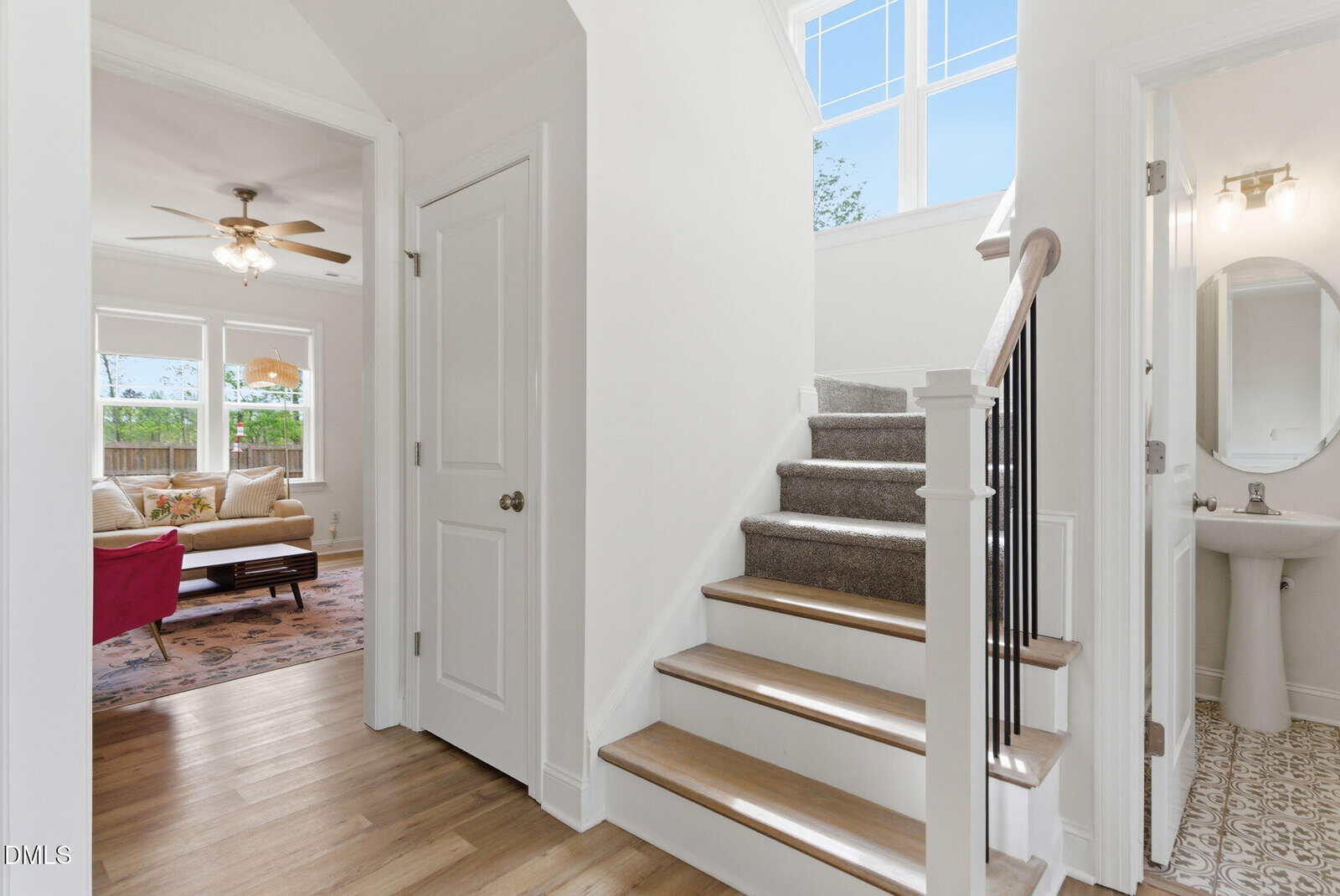 A bright entryway with light wood floors, a white door, and a carpeted staircase. A doorway leads to a living room with a couch and fan.