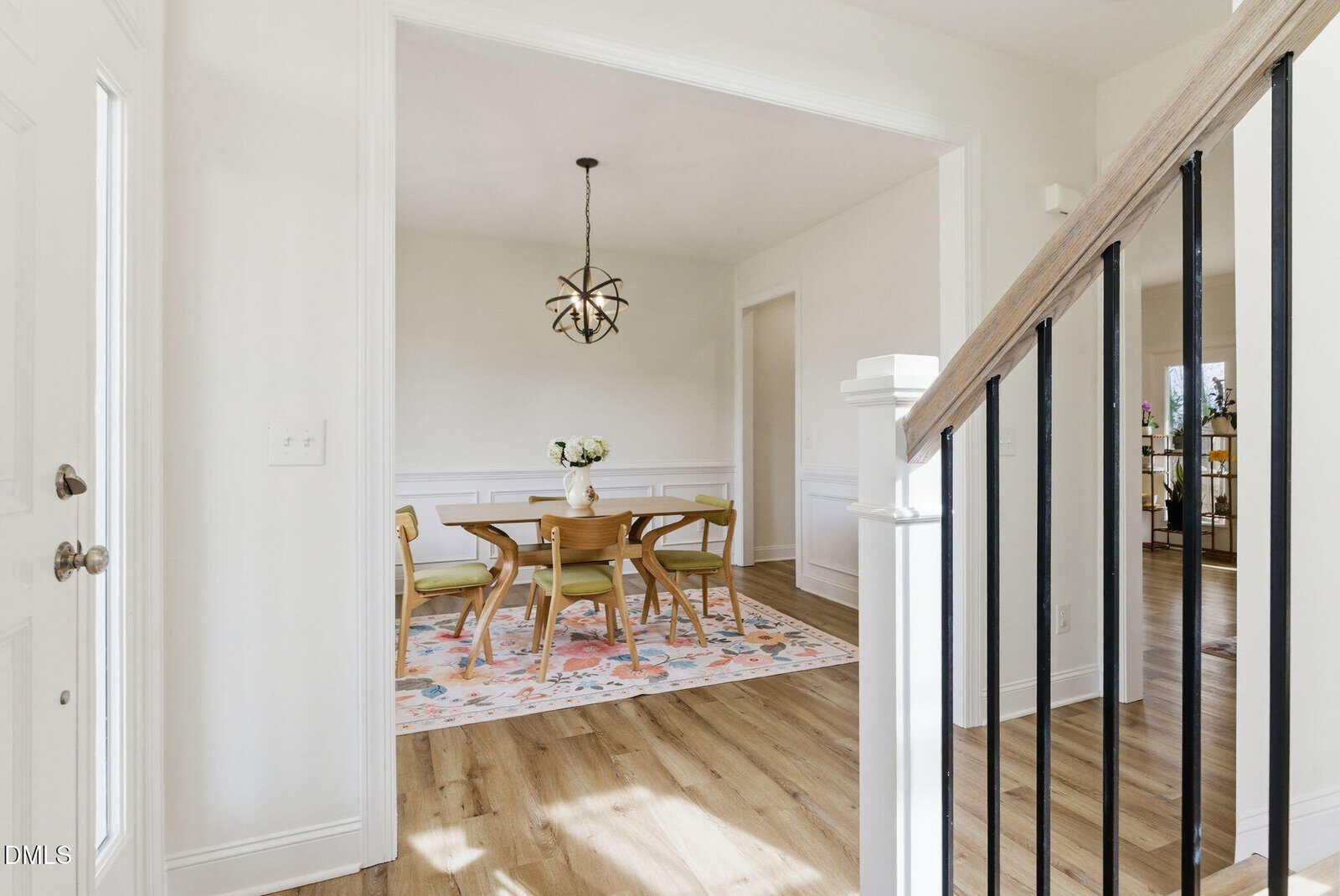 Bright entryway with staircase leading to a dining room featuring a wooden table, green chairs, floral rug, globe chandelier, and white wainscoting on hardwood floors.