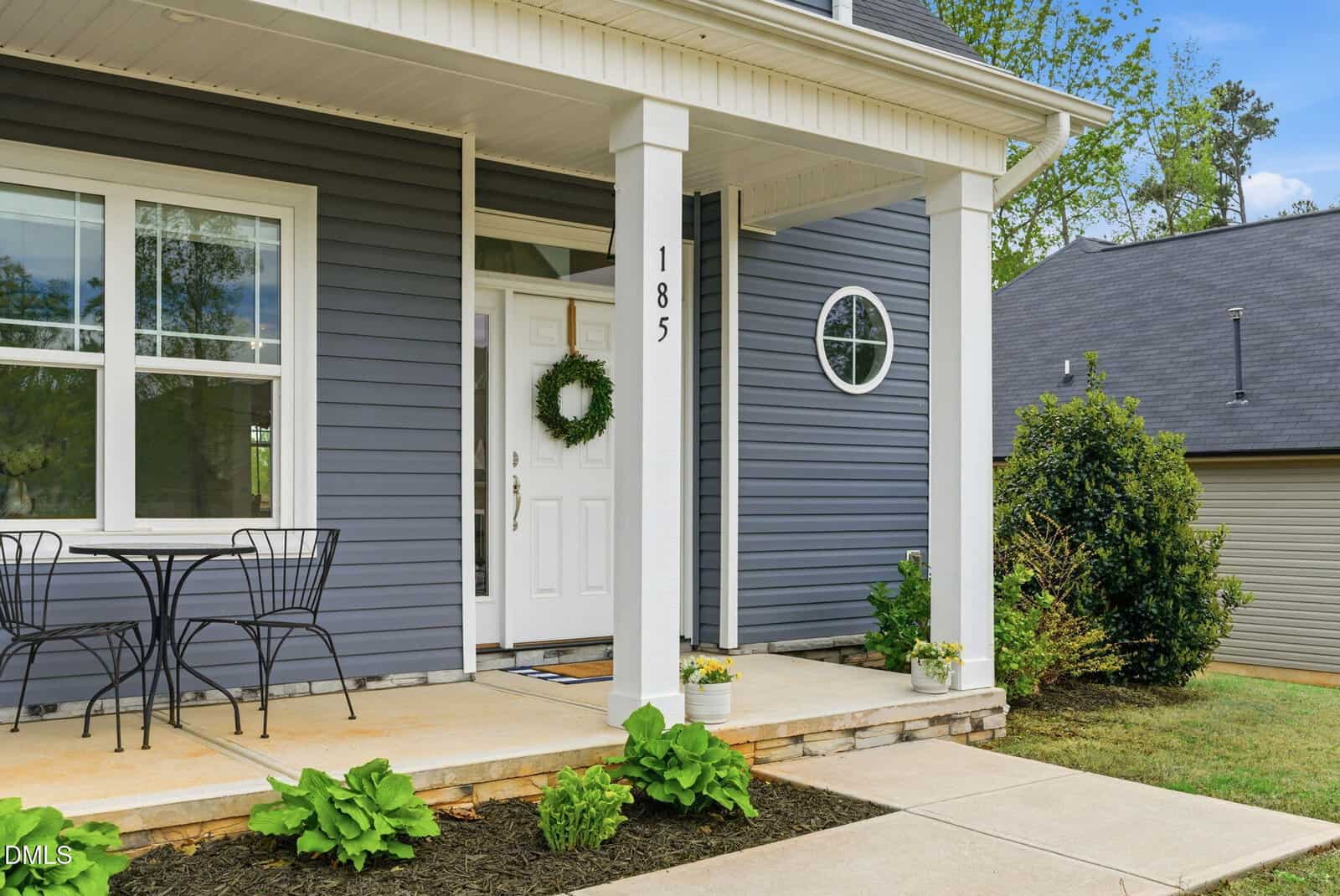 A modern house exterior with gray siding, white trim, a small front porch, white door with a green wreath, black metal bistro table and chairs, and green plants along the walkway. House number 185 is on the porch post.
