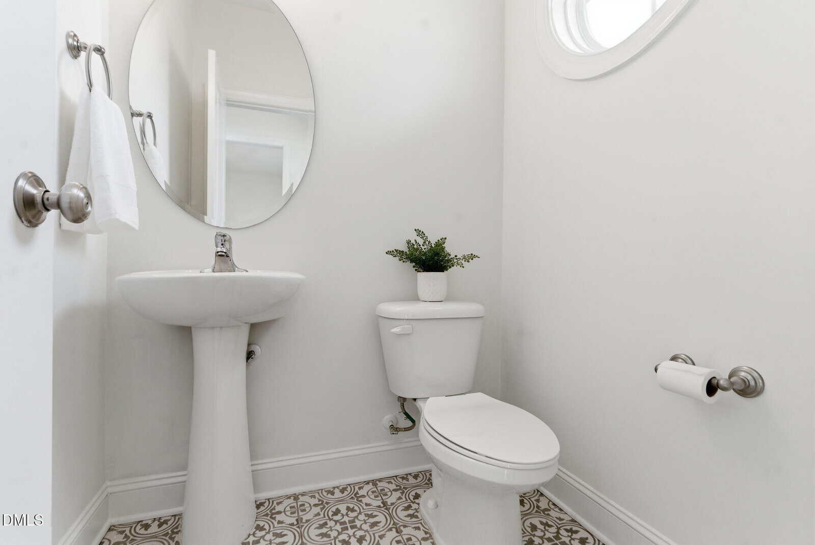 Bright powder room with pedestal sink, round mirror, white toilet with small potted plant on tank, patterned tile floor, oval window, and brushed nickel fixtures.
