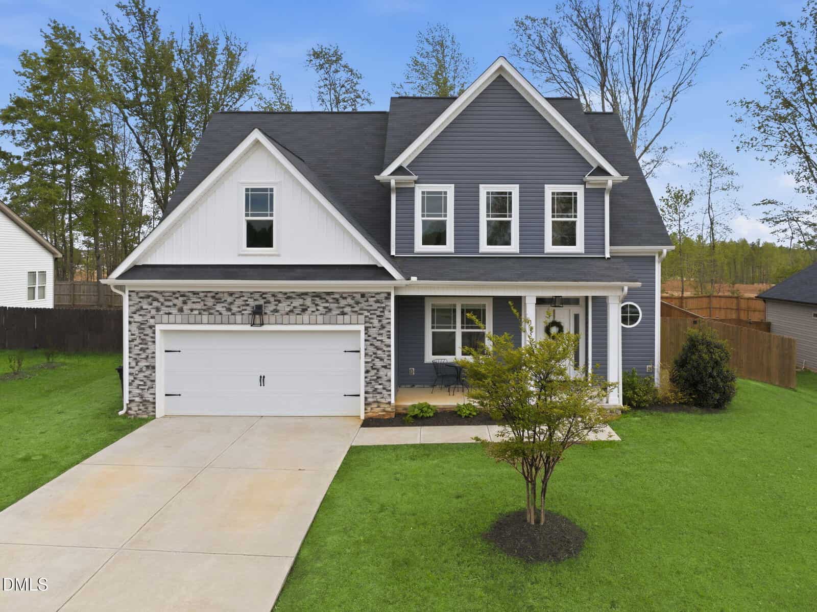 A modern two-story house with gray siding, white trim, and stone accents. It has a double garage, front porch, large windows, and a neatly manicured lawn with a small tree in front.