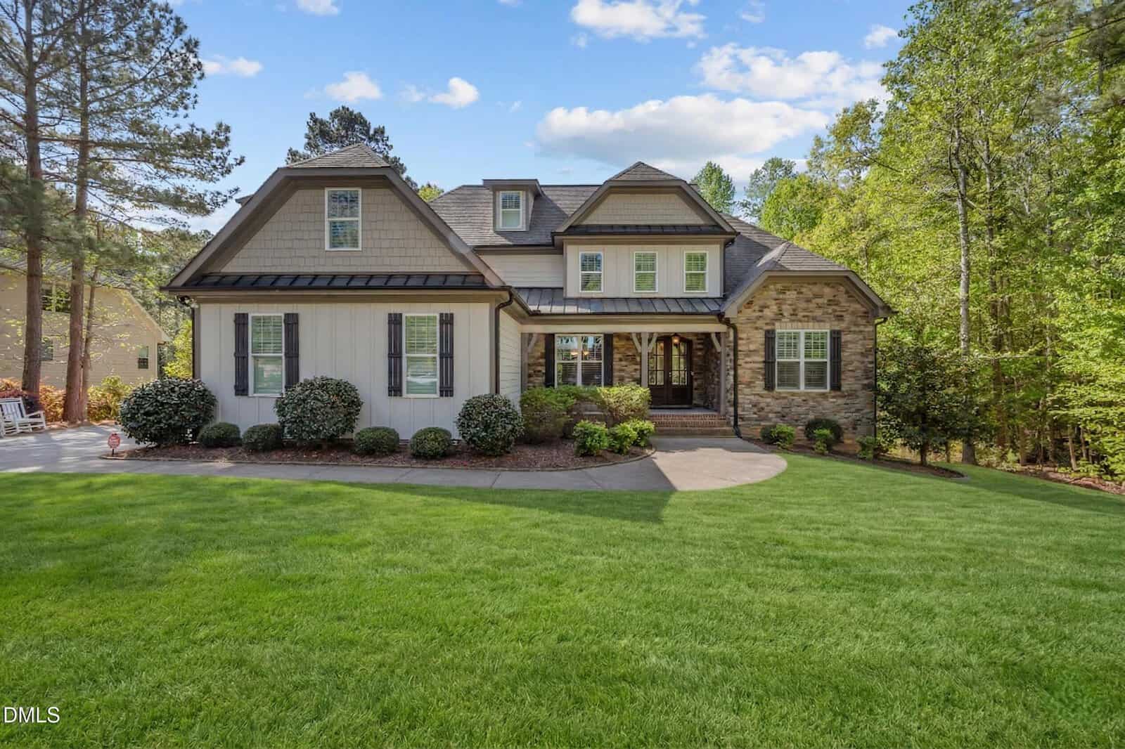 A two-story suburban house with stone and siding exterior, a covered front porch, manicured bushes, and a lush green lawn, surrounded by tall trees under a blue sky with clouds.
