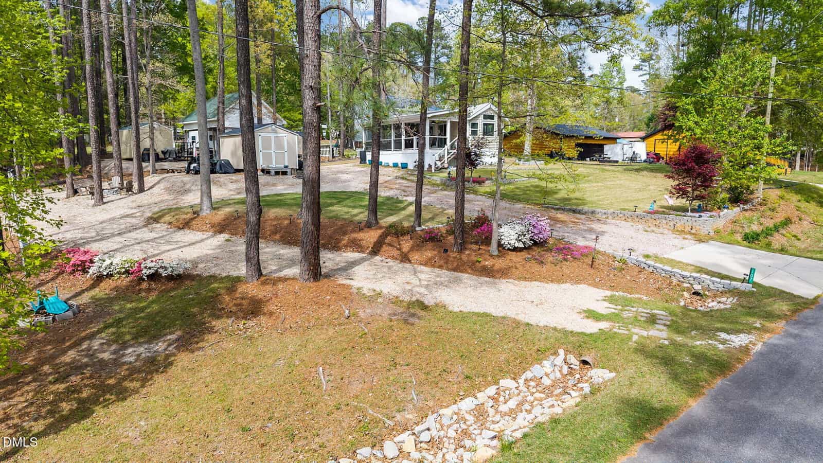 A house with a large yard and wooden shed sits among tall pine trees. A gravel driveway splits the property. Flower beds, a lawn, and a stone-edged garden appear in front, with a second building and carport to the right.