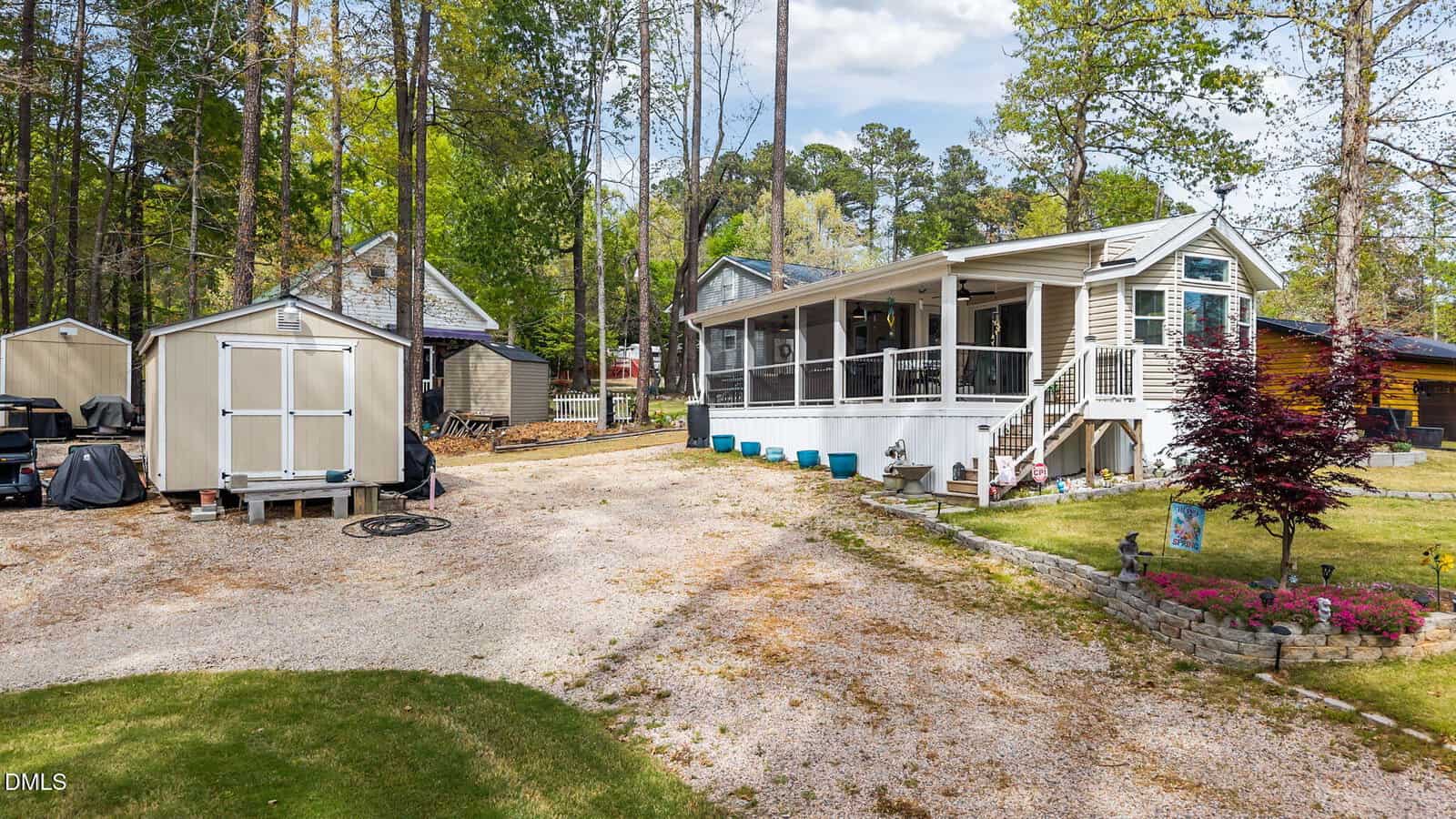 A light-colored house with a screened porch sits on a gravel driveway at 107 Lightfoot Drive in Louisburg. A white shed is to the left, surrounded by trees, with other small buildings and a landscaped yard visible in the background.