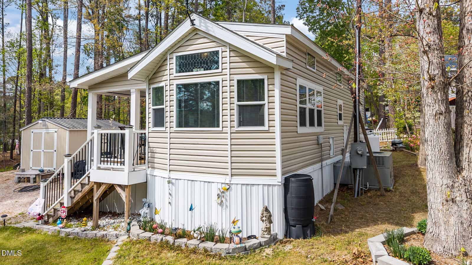 A beige mobile home with white trim sits among trees. There’s a small porch with steps, garden decorations along the front, a rain barrel, and a shed nearby. The yard is grassy and surrounded by woods.