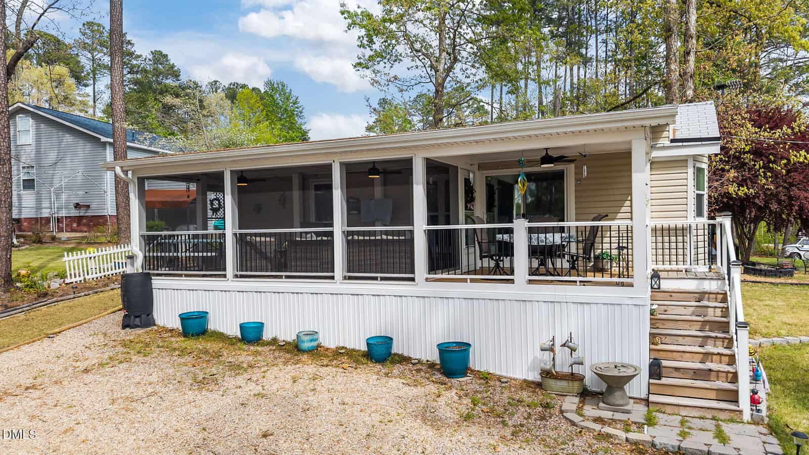 A beige house with a large screened porch, white railing, stairs, blue flowerpots, ceiling fans, and outdoor seating. Surrounded by trees, gravel, and grass. Another house is visible in the background.